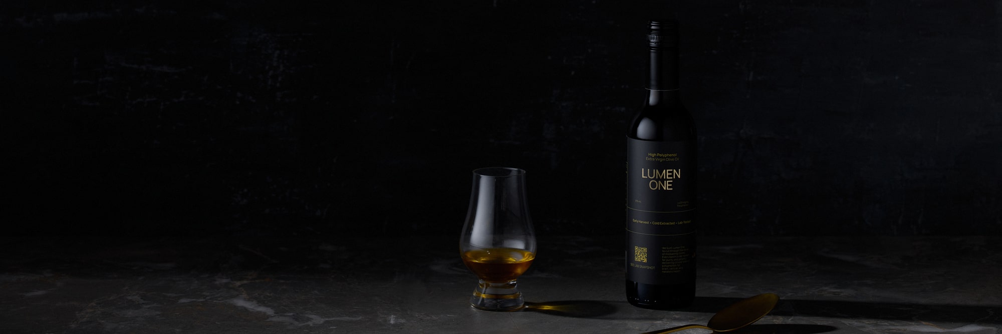 Lumen One olive oil bottle with a tasting glass and spoon on a black set, dark product photography Melbourne